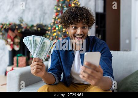 Man celebrating victory and success sitting on sofa at home in living room near Christmas tree. Hispanic man holds phone and money cash dollars in hands. A winner for the New Year holidays and Christmas. Stock Photo