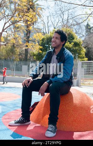 young adult afro latin venezuelan man, happy sitting in park smiling while taking care of his son when he plays, vertical image. Stock Photo