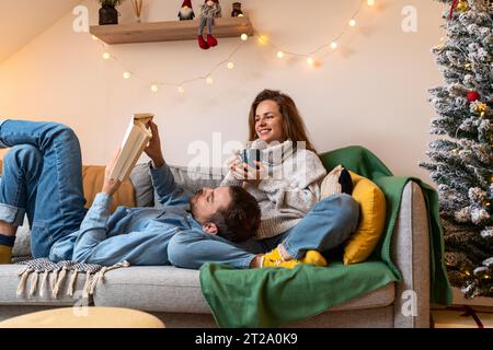Christmas holidays lifestyle. Young married couple sits on the sofa and reading book and enjoying Christmas. Stock Photo