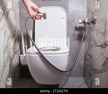 The girl holds a hygienic shower in her hand against the background of a hanging toilet bowl. The concept of care for intimate places and the preventi Stock Photo