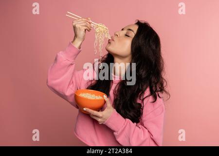 Hungry dark-haired young female snacking on tasty pasta with her eyes closed during the photo shoot on the pink background. Japanese cuisine concept Stock Photo