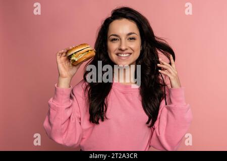 Joyous dark-haired gorgeous young woman posing on the pink background with her favorite nourishing sandwich. Fast food consumption concept Stock Photo