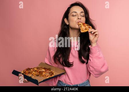 Hungry dark-haired young female eating an appetizing Italian food on the pink background during the studio photo shoot. Unhealthy nutrition concept Stock Photo