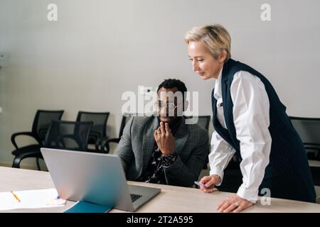 Middle-adult female boss mentor helping black male employee with online assignment teach intern in office. Businesswoman supervisor helping Stock Photo
