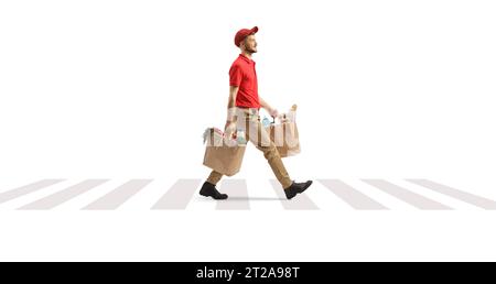 Full length profile shot of a delivery guy carrying bags with groceries and walking at a pedestrian crossing isolated on white background Stock Photo