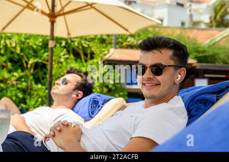 A young man in his twenties lies on a sunbed listening to music with AirPods with his brother in the background at a hotel resort in Mui Ne, Vietnam. Stock Photo