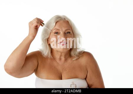 Elegant, beautiful senior woman taking care after face, applying organic serum against white studio background Stock Photo