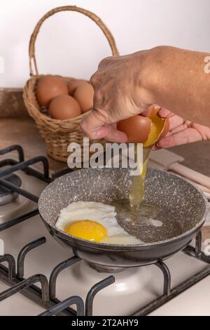 Female hands break an egg to drop it into a pan on the stove, in which another fried egg is cooking Stock Photo