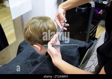 A little cute boy sits in a hairdresser's at the stylist's, a schoolchild is getting hair cut in a beauty salon, a child at a barbershop's, a short Stock Photo