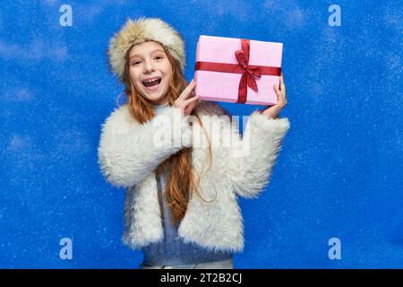 winter holidays, cheerful girl in faux fur jacket and hat holding gift box on turquoise background Stock Photo