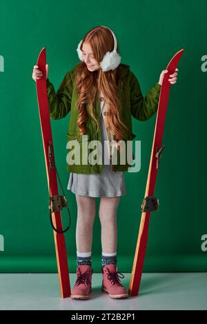 cheerful preteen girl in ear muffs and winter outfit holding red skis on turquoise background Stock Photo