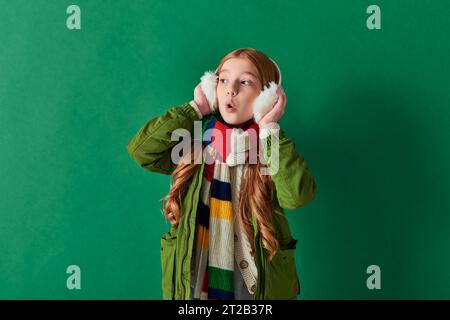 shocked preteen girl in ear muffs, striped scarf and winter outfit looking away on turquoise Stock Photo