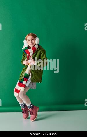 cheerful preteen girl in ear muffs, striped scarf and winter outfit posing on turquoise backdrop Stock Photo