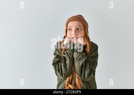 preteen girl in stylish winter outfit with knitted hat feeling cold while standing on grey backdrop Stock Photo