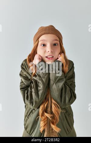 girl in winter outfit with knitted hat feeling cold and standing on grey backdrop, looking at camera Stock Photo