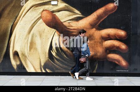 Pedestrians walking past a large reproduction of a painting by Caravaggio depicting large grabbing hand, on a hoarding in central London England UK Stock Photo