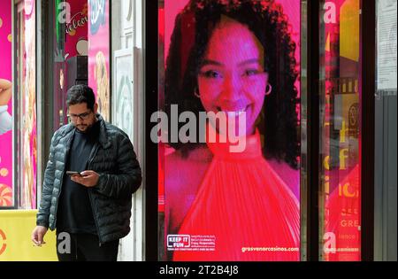 A young man using a mobile phone unaware of an electronic billboard of a smiling attractive woman, Central London England UK Stock Photo