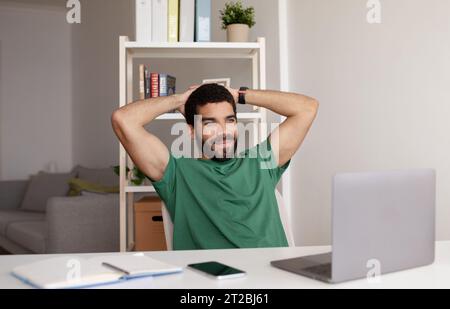 Smiling relaxed millennial arab man at table with laptop resting from work, has video call in home office Stock Photo
