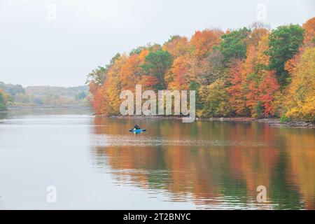 Lone fisherman in a kayak fishes near trees in beautiful autumn color on a calm overcast day Stock Photo