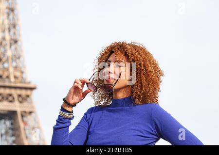 HAPPY CURLY HAIR YOUNG WOMAN IN BLUE SWEATER HOLDING HER SUNGLASSES AT THE EIFFEL TOWER Stock Photo