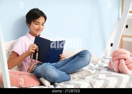 Little boy with Bible and rosary beads sitting on bed in room Stock Photo