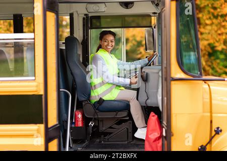 Joyful black female driver sitting in yellow school bus, smiling at camera Stock Photo