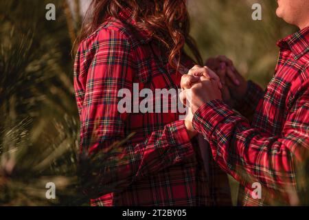 Close-up of hands, loving couple in checkered red shirts holding hands tightly among green Christmas tree market. Holiday atmosphere. Concept of tacti Stock Photo