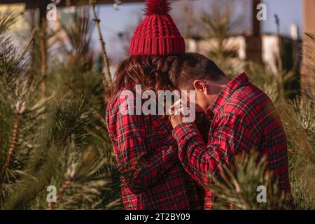 Cheerful couple in love in checkered red shirts, knitted hats are fooling around, laughing among green Christmas tree market. Young man holds hands of Stock Photo