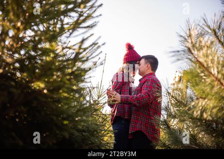 Cheerful couple in love in checkered red shirts, knitted hats are fooling around, laughing among green Christmas tree market. Young man holds hands of Stock Photo