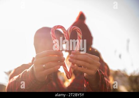 Red white candy cane is held in outstretched hands of loving couple in checkered shirts, knitted hats near green market of Christmas trees. Happy man Stock Photo