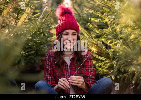 Close-up portrait of young woman with bright sparkler in hand in red plaid shirt, knitted hat among green pine trees. Curly-haired girl laughs, rejoic Stock Photo