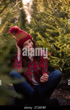 Close-up portrait of young woman with bright sparkler in hand in red plaid shirt, knitted hat among green pine trees. Curly-haired girl laughs, rejoic Stock Photo