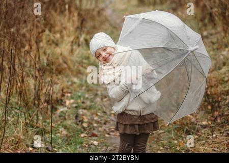 Portrait of a preschool girl standing in the park in the fall, looking out from under an umbrella, smiling. Horizontal frame. Stock Photo