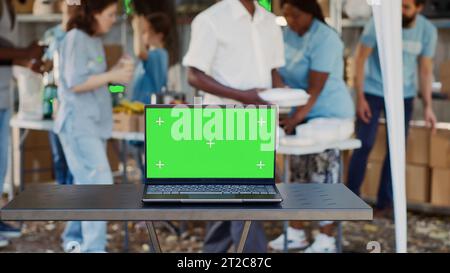 Digital laptop with blank green screen display is placed on table while volunteers help the less fortunate. Wireless computer in the foreground with isolated chromakey mockup template. Tripod shot. Stock Photo