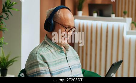 Retired traveller working on tablet in lounge area, passing time watching tv show on online streaming services. Senior man in hotel lobby using headphones and gadget, waiting to start vacation. Stock Photo