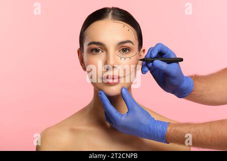 Woman preparing for cosmetic surgery, pink background. Doctor drawing markings on her face, closeup Stock Photo