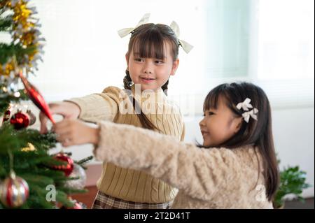 Two happy and cute young Asian girls in cute dresses are having fun and decorating a Christmas tree in the living room together. Merry Christmas, Spec Stock Photo