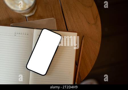 A white-screen smartphone mockup on an opened book on a wooden table in a cosy minimalist coffee shop. workspace top view Stock Photo