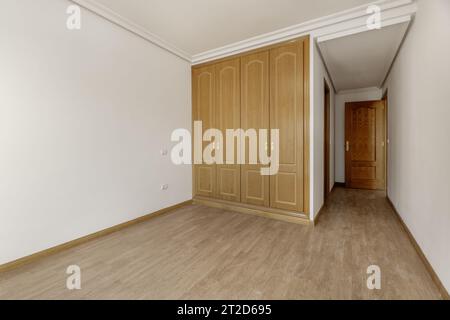 Empty bedroom with three-door built-in oak wardrobe with handles, skirting board of the same material, en-suite bathroom with wooden doors and plaster Stock Photo