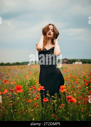 Beautiful young girl in a black evening dress posing against a poppy field on a cloudy summer day. Portrait of a female model outdoors. Rainy weather. Stock Photo