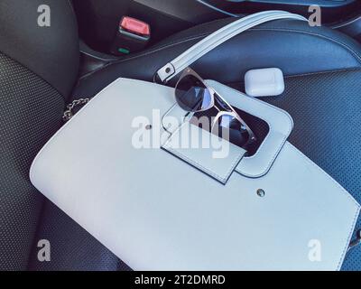White women's beautiful fashionable leather bag and sunglasses with wireless headphones lie on the leather armchair of a good expensive car. Stock Photo