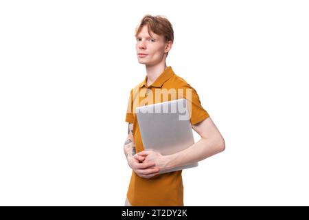 a young successful red-haired guy dressed in an orange t-shirt with a tattoo on his arm works as a programmer and holds a laptop in his hand Stock Photo