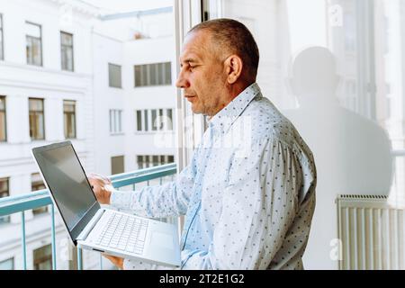 Attractive man in shirt standing near window with laptop Stock Photo
