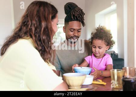 A heartwarming scene of a Caucasian mother, African father, and their daughter enjoying breakfast together in the living room, showcasing the beauty o Stock Photo
