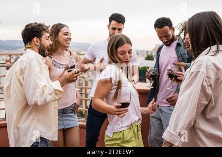 Happy young group of friends dancing in a rooftop party holding drinks.Diverse cheerful people having fun in a terrace drinking wine and listening Stock Photo