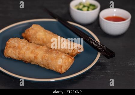 Closeup of a plate with two fried springrolls and two small bowls of dipping sauce and cucumber in the background. Stock Photo