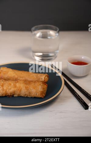 A dark blue plate with two spring rolls, a glass of water, a dipping sauce, and chopsticks on a white table, Stock Photo