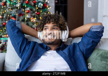 Close-up portrait of a young and smiling Muslim man sitting relaxed on the sofa at home near the Christmas tree and listening to music in headphones. Stock Photo