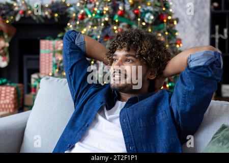 Young Indian man resting after work and study at home on sofa near Christmas tree on winter holidays. Sitting smiling and relaxed on the couch. Close-up photo. Stock Photo
