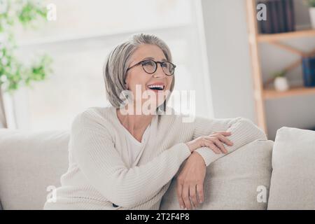 Photo of good mood thoughtful elderly lady wear white cardigan spectacles having rest couch smiling indoors apartment room Stock Photo